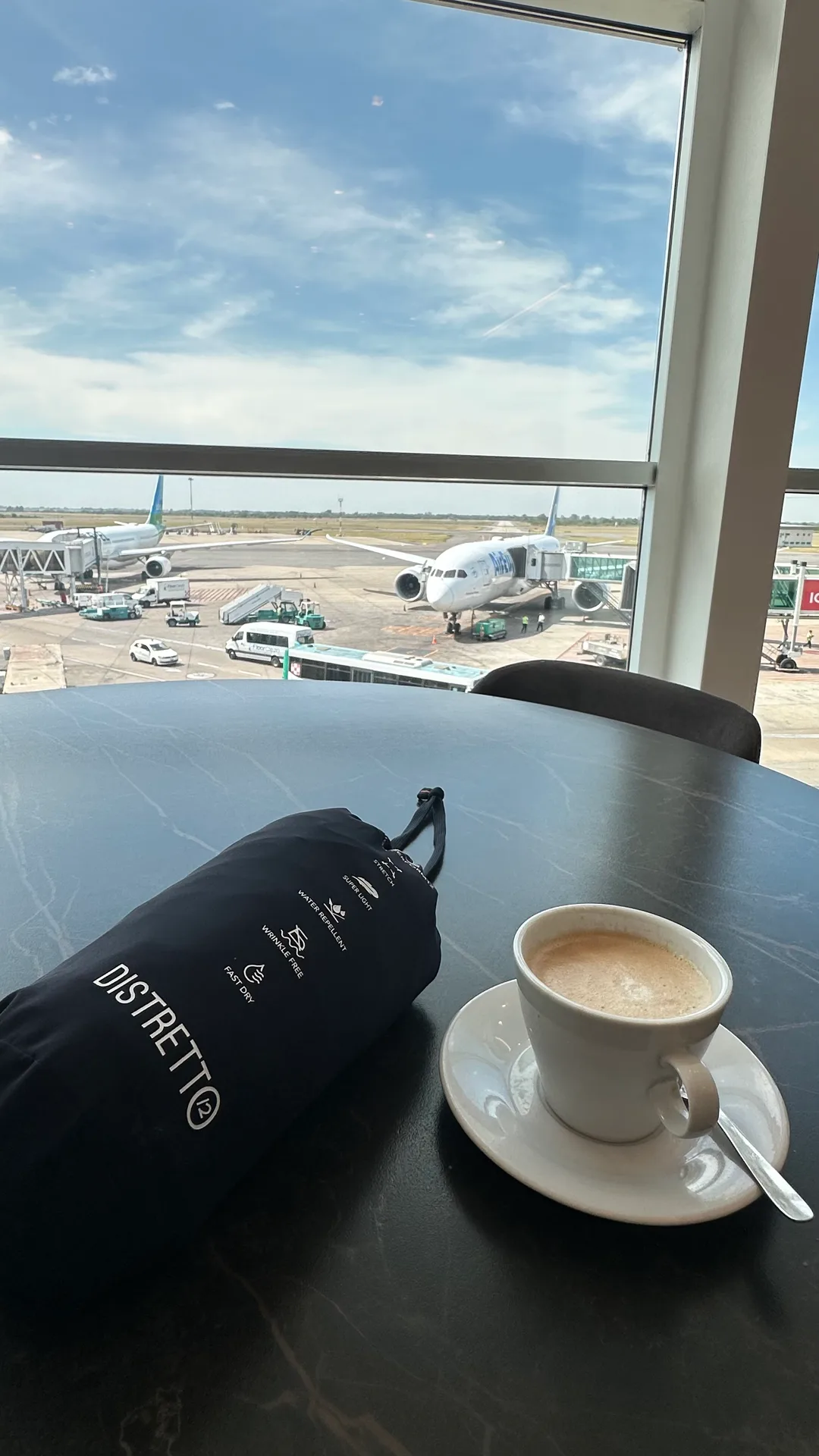Bolsa de viaje de Distretto12 sobre una mesa de aeropuerto, con un café y un avión de fondo